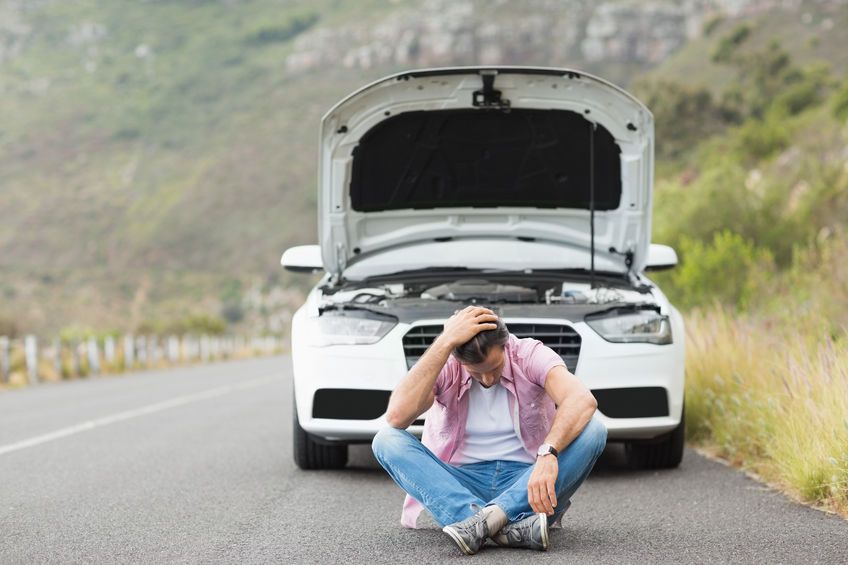 A car broken down on the side of the road with the driver sitting on the pavement, distressed.